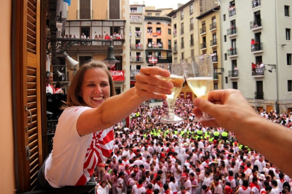 Pamplona Running of the Bulls 2026: Complete Travel Guide & Cultural Context photo | Cosmopolitours Woman smiling and toasting with champagne from a private balcony during the San Fermín festival in Pamplona, overlooking the packed crowd in festive white and red below.