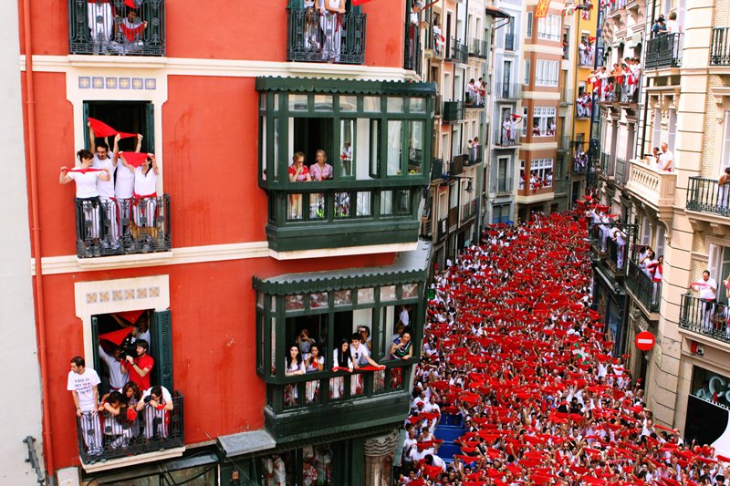 Pamplona Running of the Bulls 2026: Complete Travel Guide & Cultural Context photo | Cosmopolitours Crowd of festival-goers in red and white fills the streets of Pamplona during San Fermín’s opening ceremony, with locals and visitors watching from historic balconies.