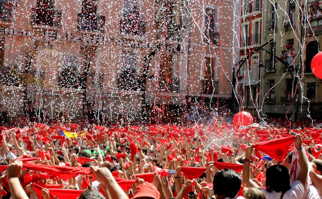Inside the Running of the Bulls: A Complete Cultural Guide to Pamplona’s San Fermín Festival photo | Cosmopolitours Crowd of people holding red scarves in the air during the Chupinazo opening ceremony of the San Fermín Festival in Pamplona, with confetti raining down and balconies filled with spectators.