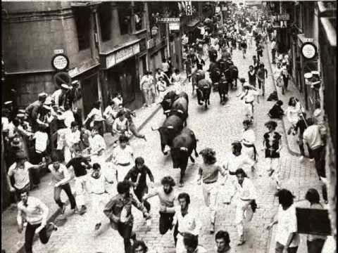 Inside the Running of the Bulls: A Complete Cultural Guide to Pamplona’s San Fermín Festival photo | Cosmopolitours Black-and-white photo of the Running of the Bulls in Pamplona, showing a crowd of runners ahead of a group of bulls charging through narrow cobblestone streets, with spectators watching from balconies.