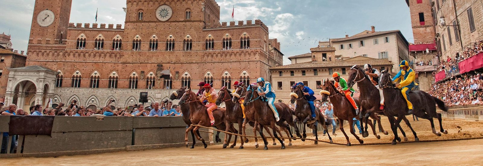 Bareback jockeys racing at full speed around Piazza del Campo during the Palio di Siena, with crowds and Siena’s Palazzo Pubblico in the background.